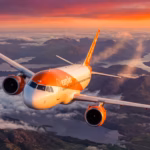 An easyJet A320 Neo flying above clouds and under an orange, red and yellow sunset. The aircraft is flying over a body of water and land.