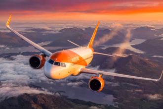 An easyJet A320 Neo flying above clouds and under an orange, red and yellow sunset. The aircraft is flying over a body of water and land.