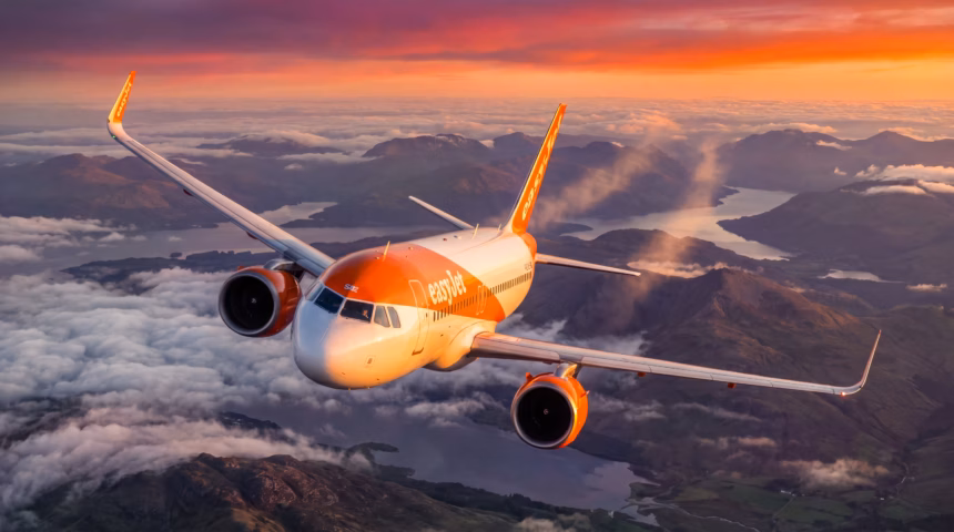 An easyJet A320 Neo flying above clouds and under an orange, red and yellow sunset. The aircraft is flying over a body of water and land.