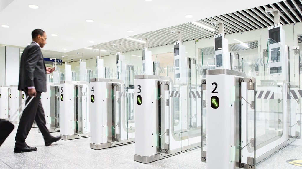 eGates shown inside Bristol Airport. White interior with yellow arrows pointing which way to walk through the system. A man with a suitcase awaiting his turn.