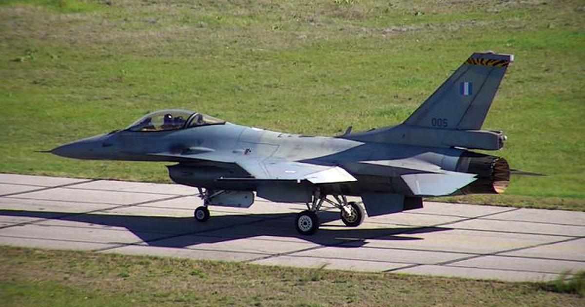 The Hellenic Air Force F-16 jet in a grey colour with the greek flag on the side, taxiing at an airport runway.