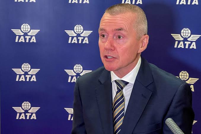 William Walsh wearing a blue suit and striped tie standing in front of am IATA wall.