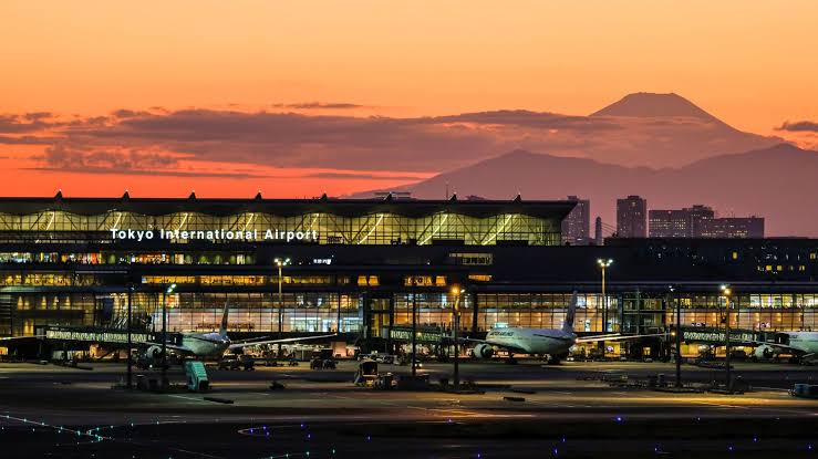HND entrance at sunset with a mountain in the background. cars are parked in front of the building which has lots of lights.