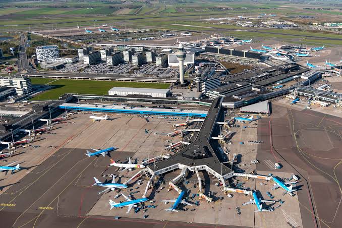 The outside of Schipol airport. One can see several aircraft parked outside of the gates. Behind the terminal, there is a large field of grass.