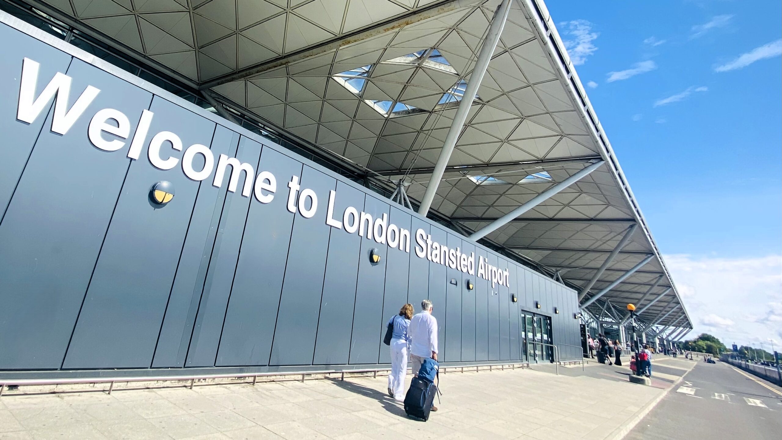 Exterior view of London Stansted Airport terminal, featuring a large wall-mounted sign reading “Welcome to London Stansted Airport.” Travelers walk along the pavement outside the terminal.