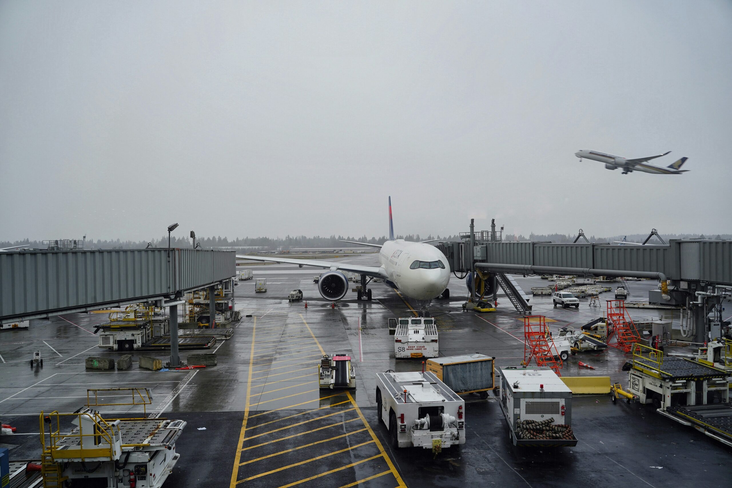 A passenger aircraft parked at an airport gate on a rainy day, surrounded by jet bridges, ground service vehicles and equipment. In the background, another airliner is taking off beneath a grey, overcast sky