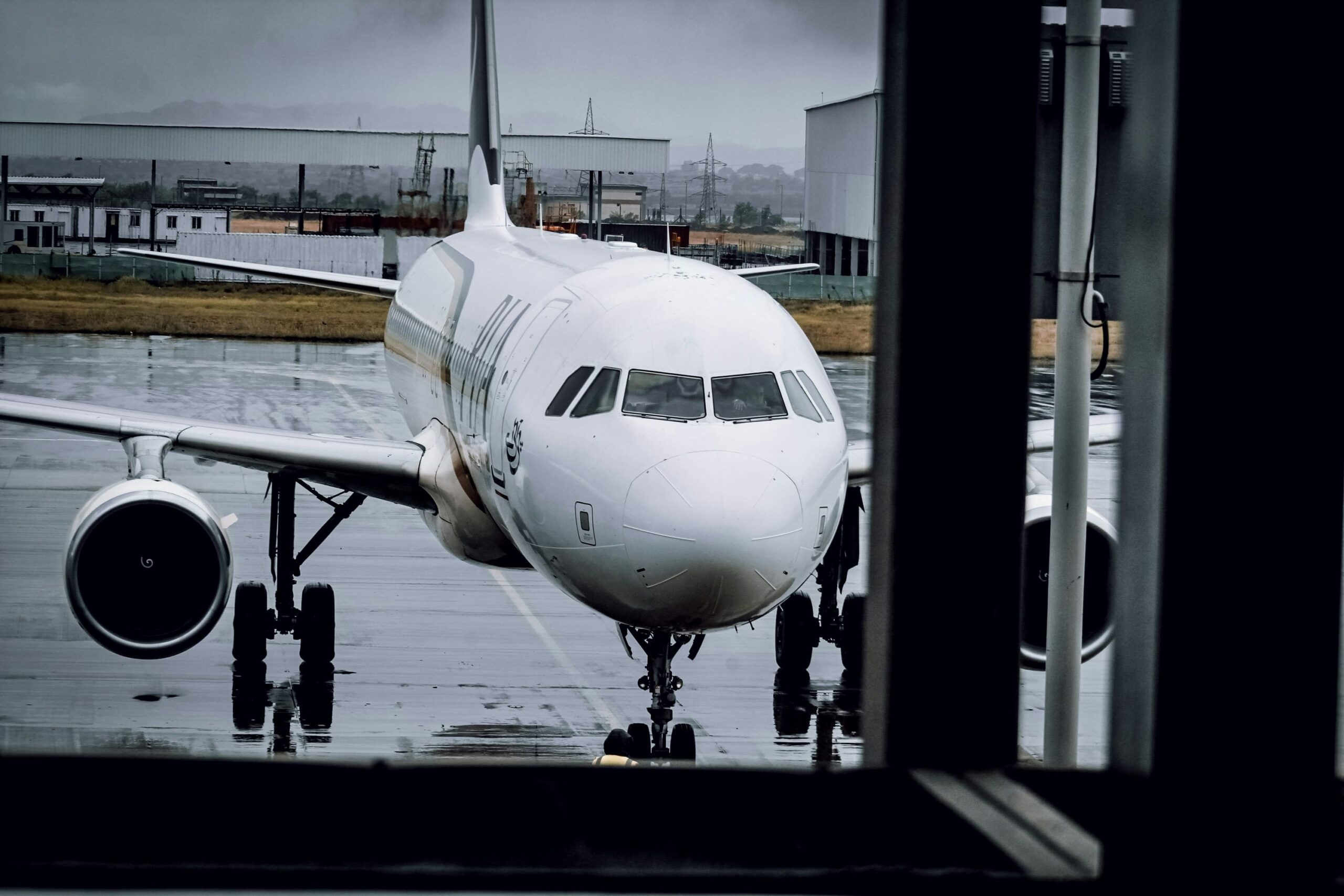 A large Airbus A320 jetliner grounded at Islamabad Airport, sitting on top of airport tarmac. The picture is taken from inside a building looking out at the front of the plane, and the right side is partly obscured by the edge of the widow. The aircraft is white, and the branding for Pakistan International Airlines can just about be seen across the side of the fuselage. The ground appears wet and the sky is grey, foggy, and cloudy in the background.