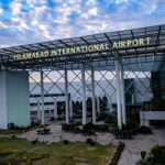 An entrance building at Islamabad Airport. In the foreground is a path surrounded by grass and close to the ground foliage leading up to the rectangular arch adorning the entrance. A sign on the arch reads 'ISLAMABAD INTERNATIONAL AIRPORT'. In the background a blue sky with white clouds can be seen.