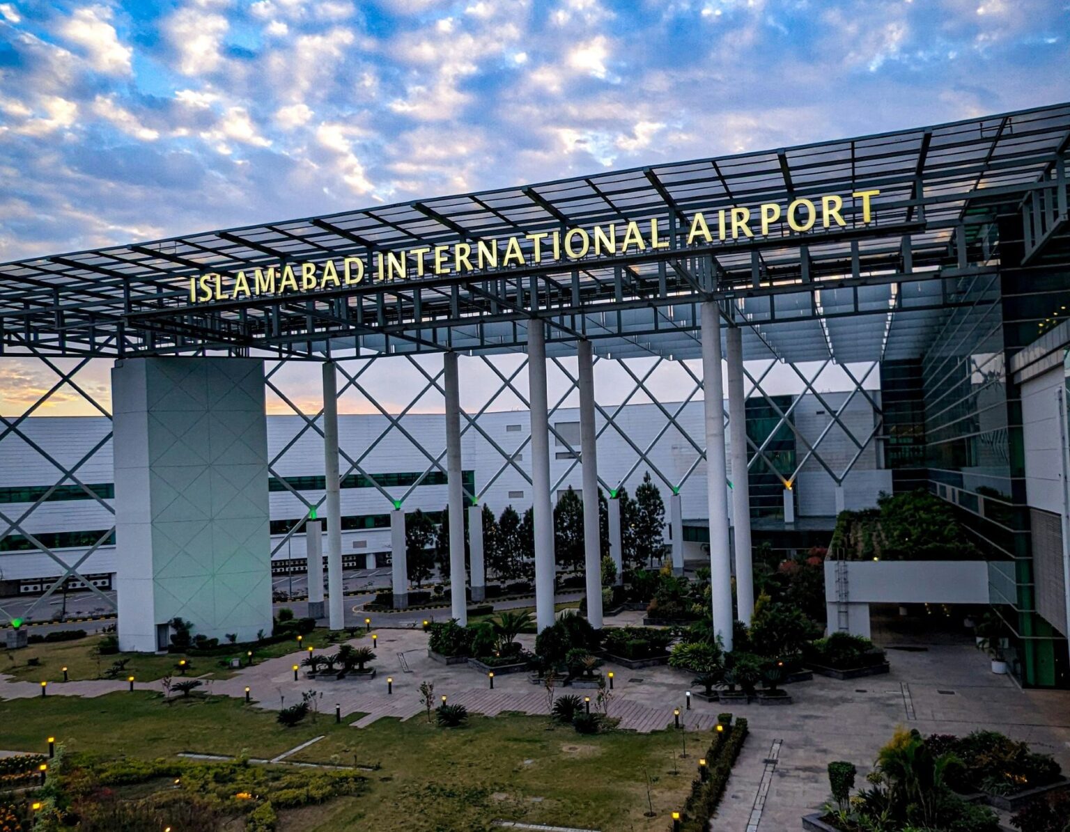 An entrance building at Islamabad Airport. In the foreground is a path surrounded by grass and close to the ground foliage leading up to the rectangular arch adorning the entrance. A sign on the arch reads 'ISLAMABAD INTERNATIONAL AIRPORT'. In the background a blue sky with white clouds can be seen.
