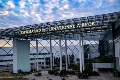 An entrance building at Islamabad Airport. In the foreground is a path surrounded by grass and close to the ground foliage leading up to the rectangular arch adorning the entrance. A sign on the arch reads 'ISLAMABAD INTERNATIONAL AIRPORT'. In the background a blue sky with white clouds can be seen.