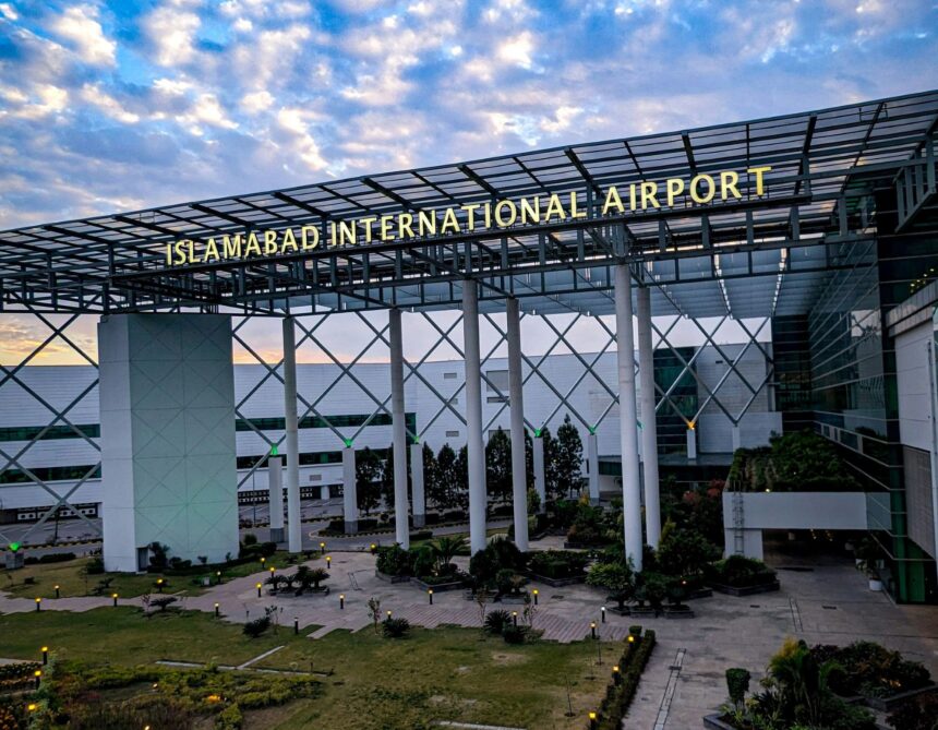 An entrance building at Islamabad Airport. In the foreground is a path surrounded by grass and close to the ground foliage leading up to the rectangular arch adorning the entrance. A sign on the arch reads 'ISLAMABAD INTERNATIONAL AIRPORT'. In the background a blue sky with white clouds can be seen.