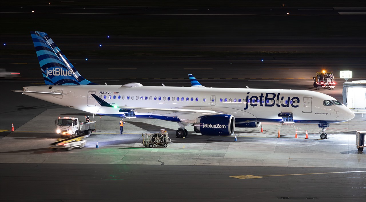 JetBlue Airbus A220-300 registered N3169J parked at a gate at night with ground handling equipment nearby