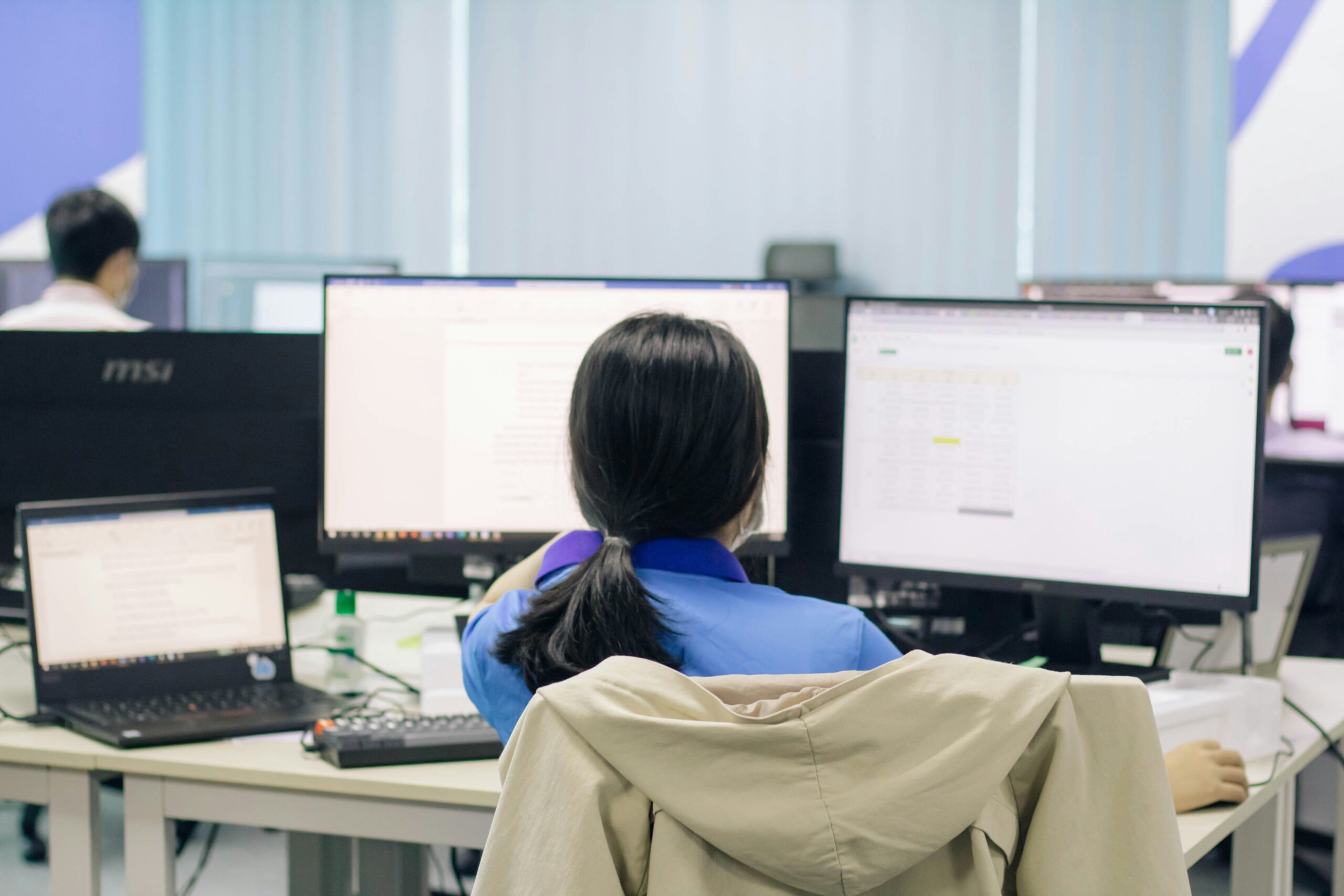 A generic image of a person with long black hair tied back into a low ponytail is pictured sat in front of multiple computer screens. The person is wearing a light blue top, and there is a beige jacket hung over the back of their chair. The screens are stylistically blurred but eminate a white light. The backgroun dis also stylistically blurred, but the pictured scene appears to be inside an office like setting. 