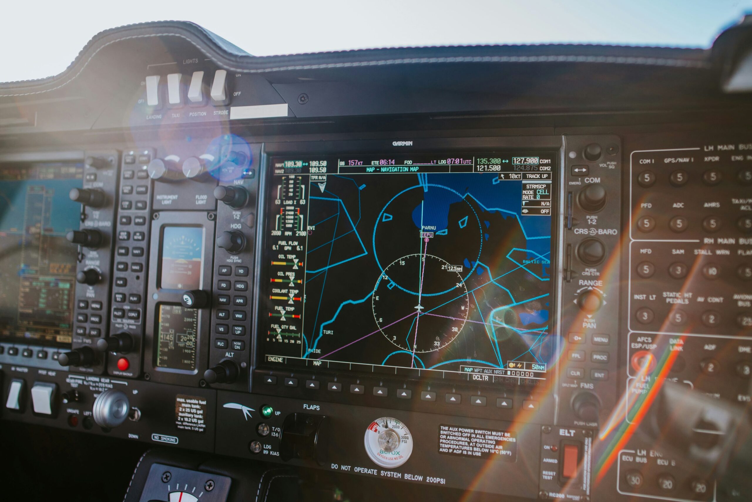A the dashboard controls in an aircraft. The hardware is black. Two screens are can be seen with maps and navigation displays visible. Sunlight beams into the cockpit from the windscreen. 
