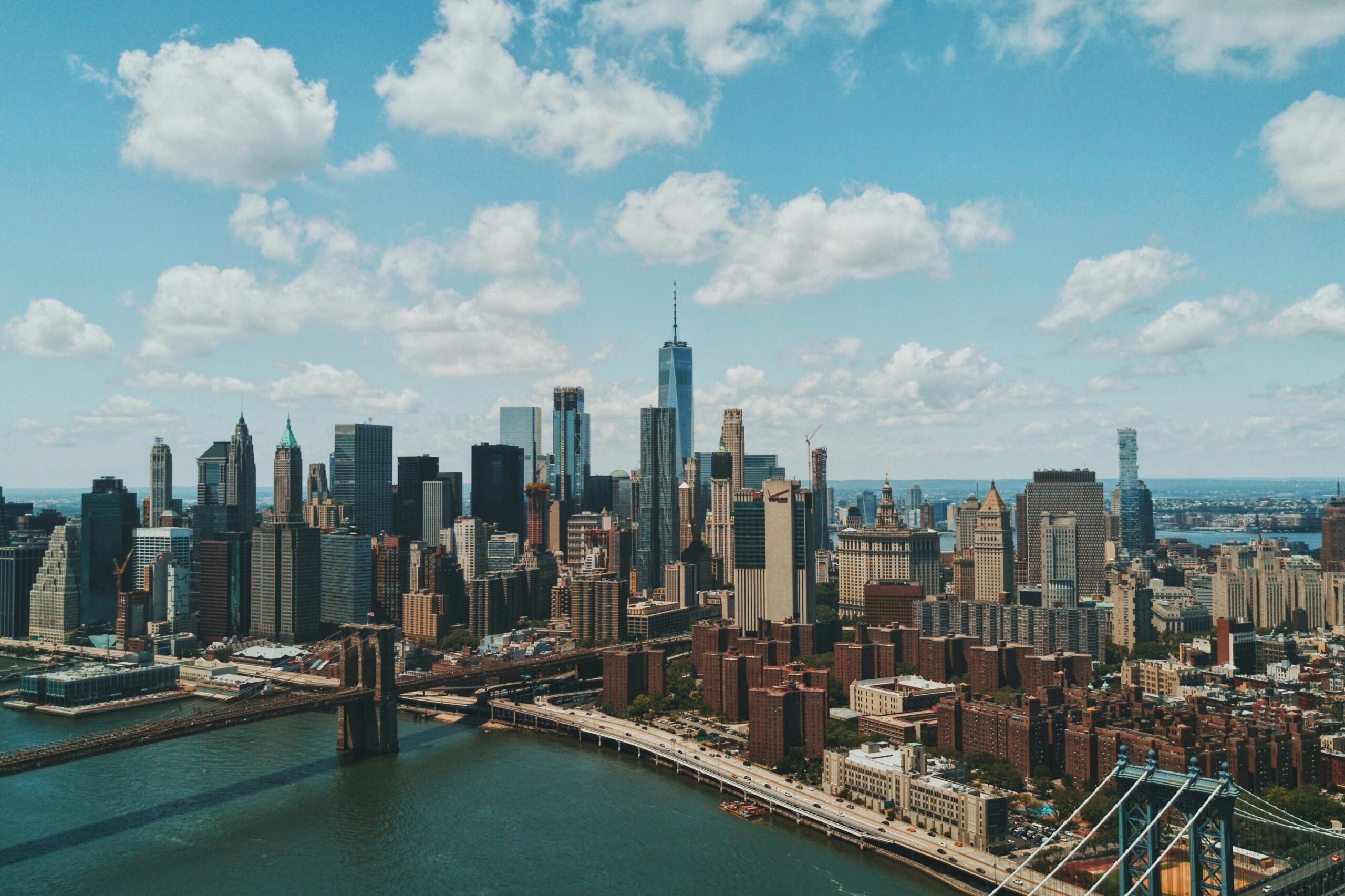 A city skyline of tall skyscrapers and high-rise buildings beside a river, with bridges crossing the water in the foreground. The scene is viewed from above under a bright blue sky with scattered clouds