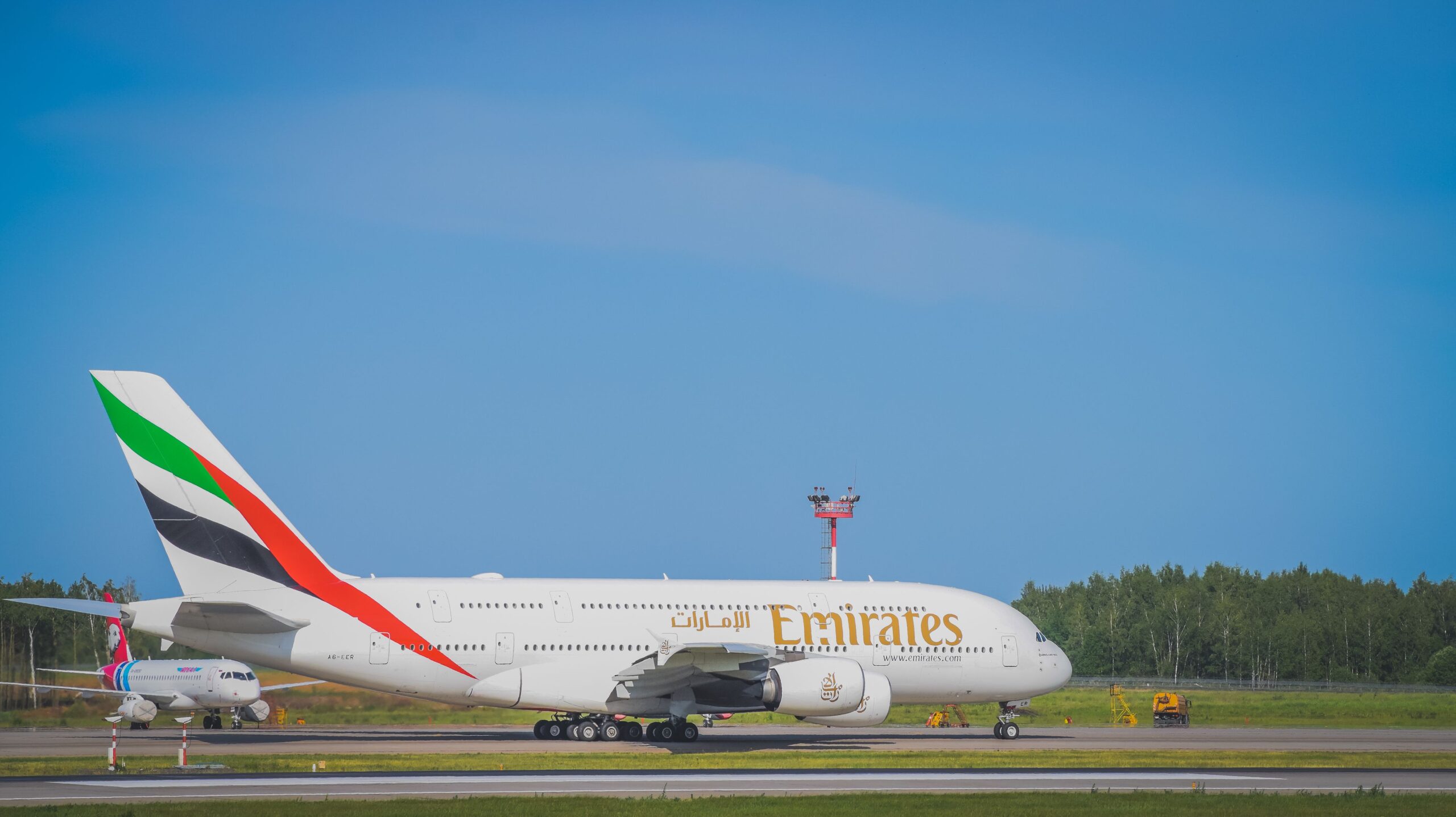 Emirates Airbus A380 taxiing on a runway, with the airline’s gold branding and UAE flag tail livery visible. A smaller passenger aircraft and airport infrastructure can be seen in the background under a clear blue sky