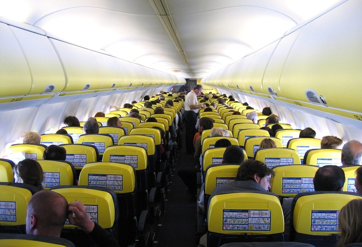 The interior of a modern plane taken from the back of the cabin, showing the backs of rows of seats and all the way down the aisle.