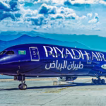A Riyadh Air dark blue B787-9 standing in front of mountains while parked in a runway. The sky is blue with clouds.