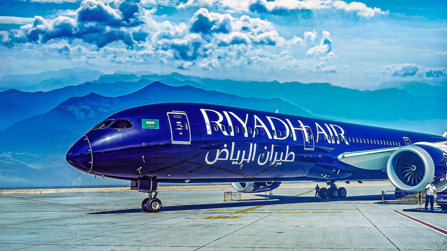 A Riyadh Air dark blue B787-9 standing in front of mountains while parked in a runway. The sky is blue with clouds.