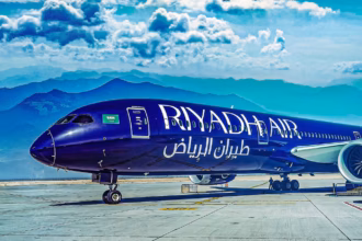 A Riyadh Air dark blue B787-9 standing in front of mountains while parked in a runway. The sky is blue with clouds.