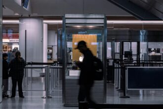 A couple silhouettes of people can be seen walking through and past border security at an airport. It is a shutter-speed style photo, stylistically blurred.