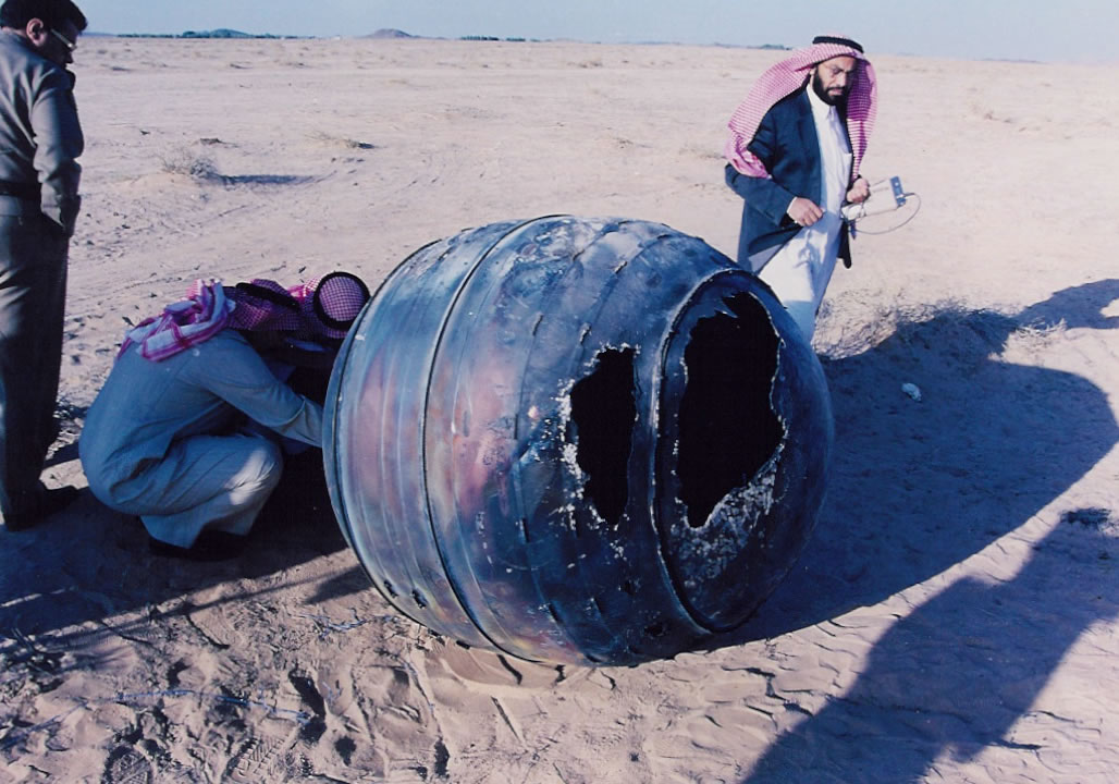 The blackened and damaged spherical titanium motor casing of a Delta II rocket, weighing about 70 kg, lands in Saudi Arabia. The debris is being inspected by four men.