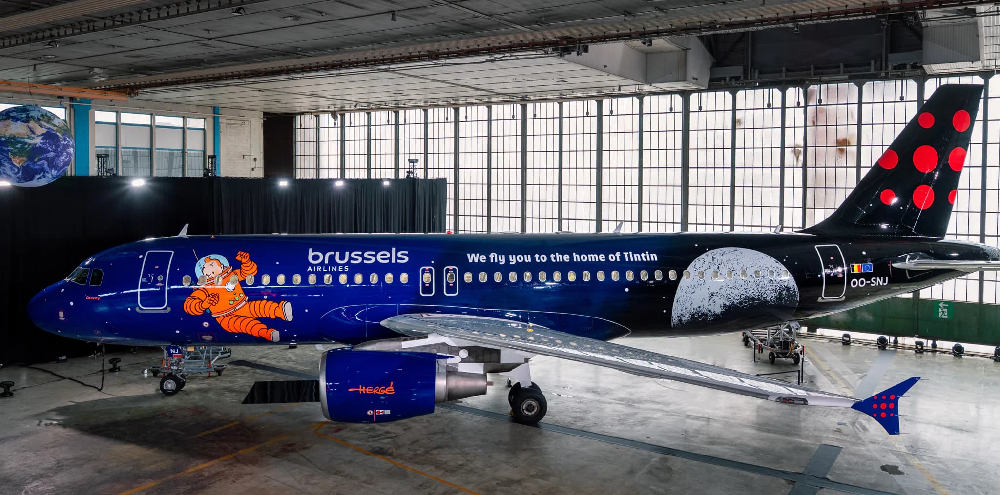 Airbus A330 with Tintin icon on the side, in a dark navy blue with moon- writing "we fly you to the home of Tintin" aircraft is in the hanger
