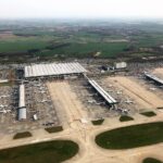 An aerial view of London Stansted Airport where the tarmac situates many stationary aircraft. A part of the airport is visible with the sky seemingly clear.