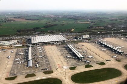 An aerial view of London Stansted Airport where the tarmac situates many stationary aircraft. A part of the airport is visible with the sky seemingly clear.