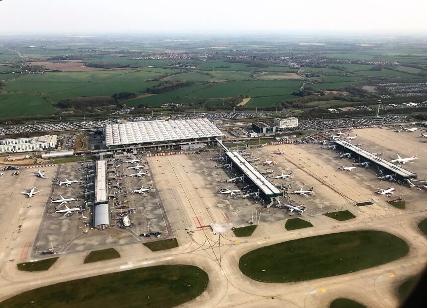 An aerial view of London Stansted Airport where the tarmac situates many stationary aircraft. A part of the airport is visible with the sky seemingly clear.