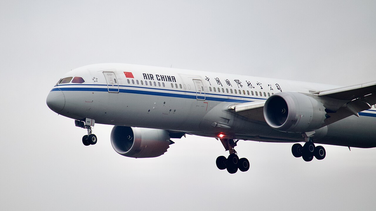 A white with two blue stripes Air China Boeing 787 plane landing at Frankfurt Airport. In the background the sky appears cloudy and grey. The aircraft has two jet engines and all wheels are out as it readies to land.