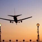 An aircraft landing at Heathrow Airport. The sun is setting creating a ombre of blue and orange. Run way lights can be seen in the forefront of the photo as the plane prepares to land.