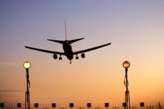An aircraft landing at Heathrow Airport. The sun is setting creating a ombre of blue and orange. Run way lights can be seen in the forefront of the photo as the plane prepares to land.