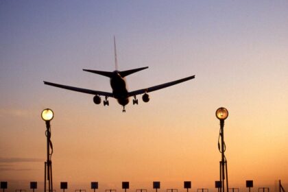 An aircraft landing at Heathrow Airport. The sun is setting creating a ombre of blue and orange. Run way lights can be seen in the forefront of the photo as the plane prepares to land.