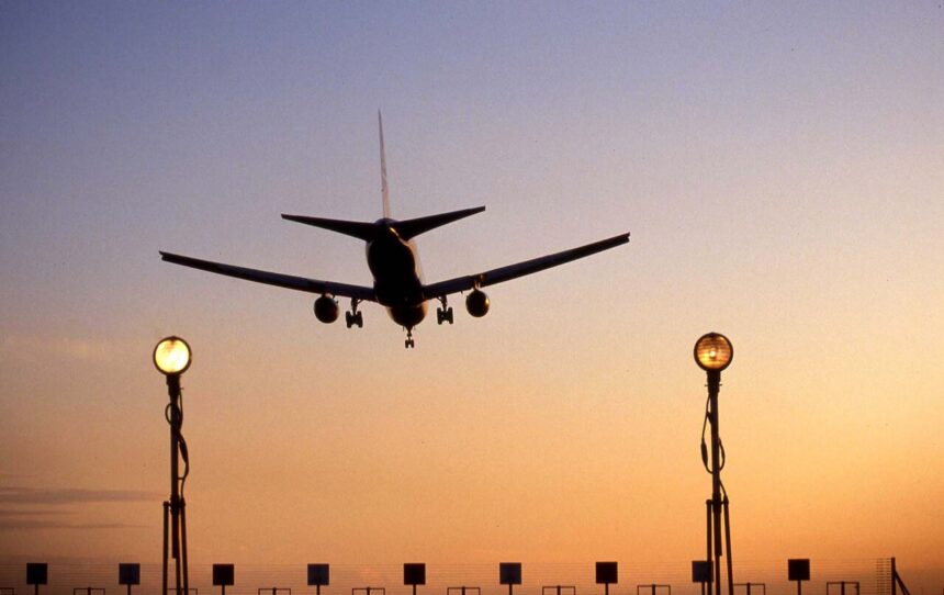 An aircraft landing at Heathrow Airport. The sun is setting creating a ombre of blue and orange. Run way lights can be seen in the forefront of the photo as the plane prepares to land.