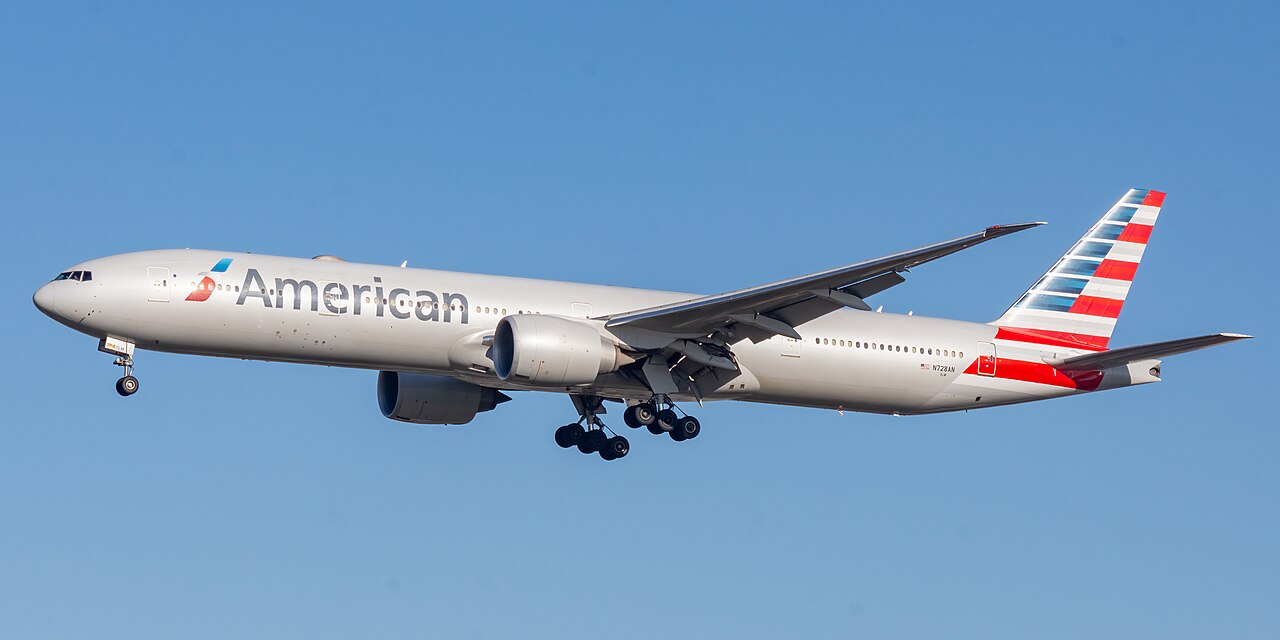 An American Airlines B777 flying under a blue sky. The aircraft's liver is mostly white with its tail displaying the US' flag.