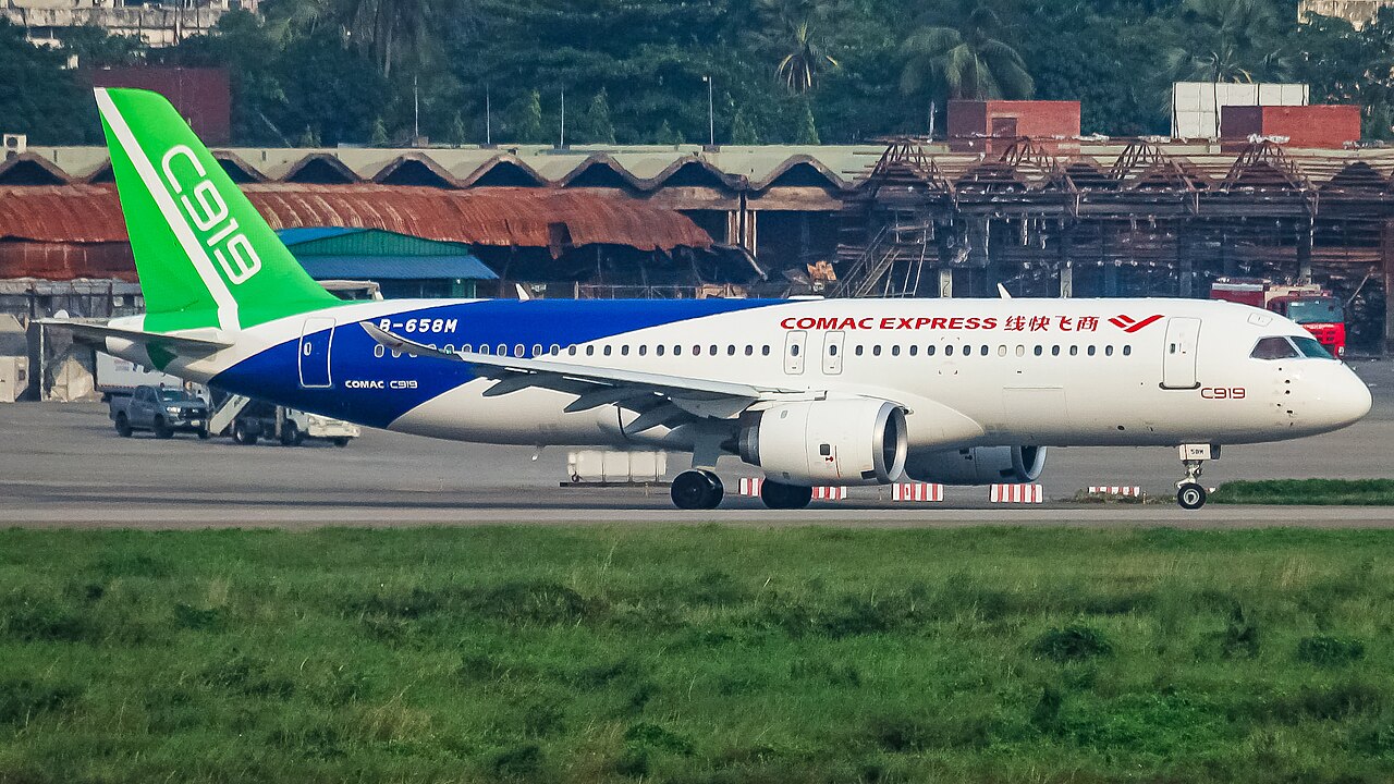 A COMAC C919 airplane on the runway. The planes body is white with bright blue and green accents at the tail of the aircraft, along with red font that says COMAC express. In the background is an unbusy airport runway.