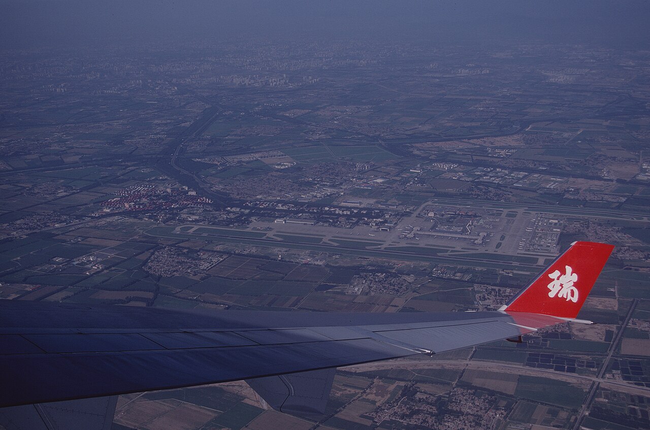 Cloudy aerial view of Beijing Capital International Airport with the wing of an airplane partially in view. The wing has a Chinese symbol in white on a bright red wingtip.