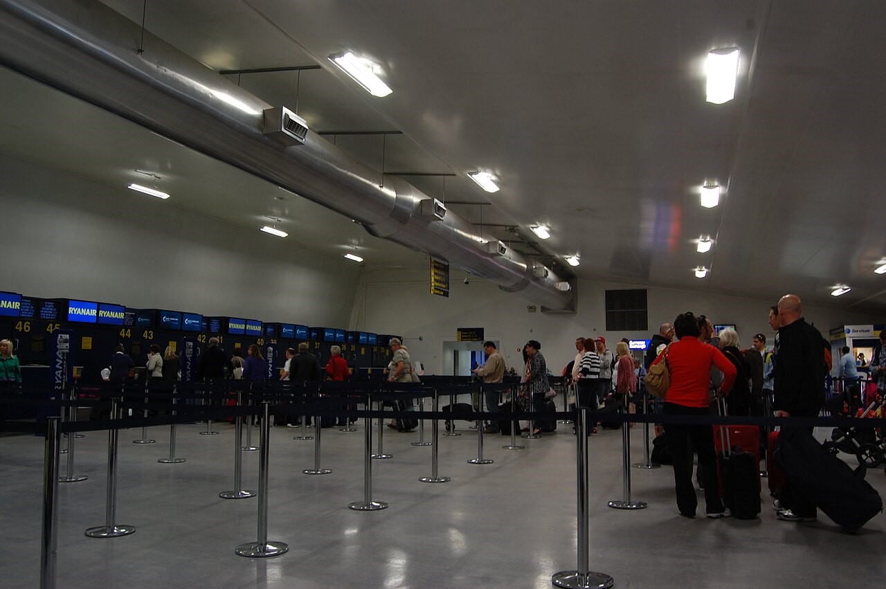 The inside of East Midlands Airport departure hall where people are queuing up to depart. People are stood with suitcases in a small line that doesn't appear overly busy. The hall is white with bright lights.