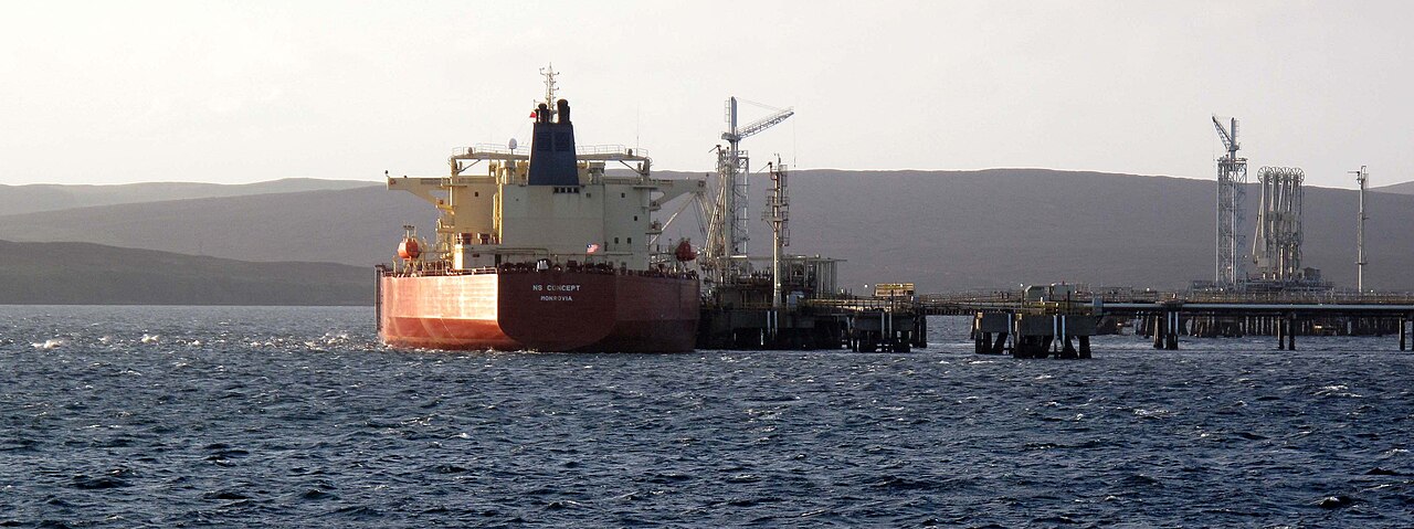 A massive ship that is loading Brent Crude oil from a oil rig to supply it to elsewhere. The ocean looks still from where the ship is anchored, and the sky is overcast in the background.