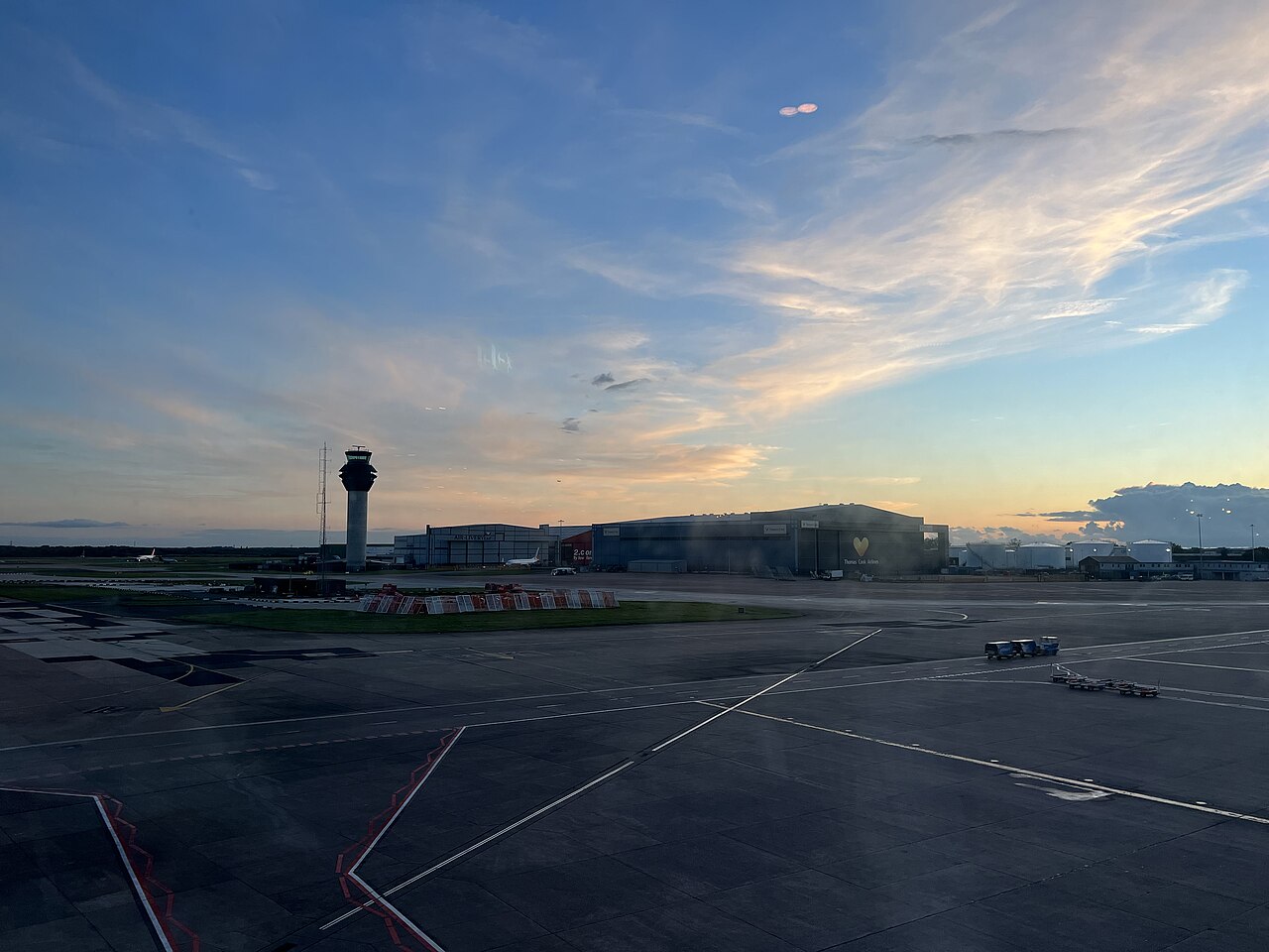 Manchester Airport at dusk, the sky is shades of blue and orange with wispy clouds above the runway. The sun has just sunk below the skyline and Manchester Airport's control tower stands in the distance.