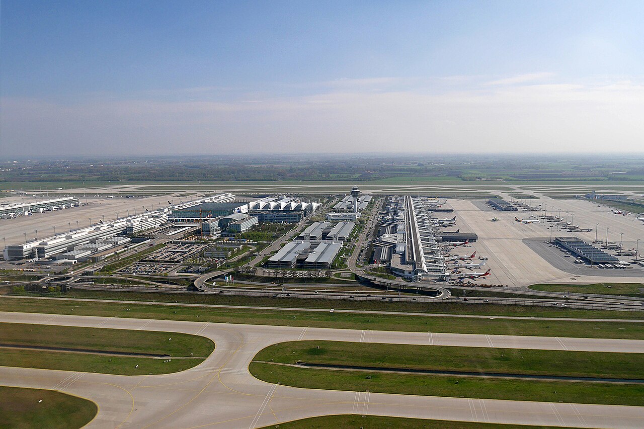 Aerial view of Munich Airport with Terminal 1, Terminal 2 and Munich Airport Center. The sky is blue and clear with green grass surrounding the airport runway.