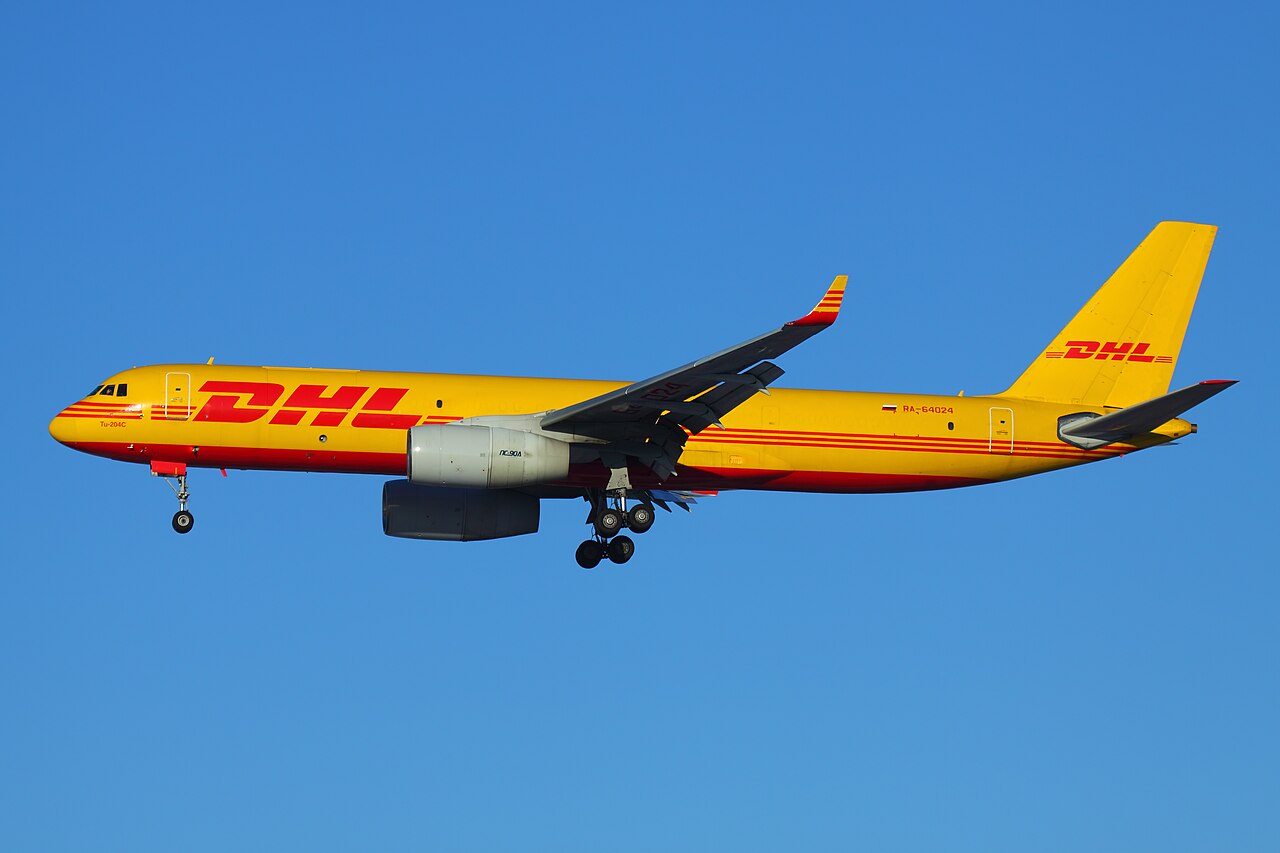A DHL airplane in mid-flight. It's yellow and red colour theme completely decorate the plane and stand out against the bright blue sky in the background.
