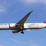 A Turkish Airlines B787-9 plane mid flight. The plane is decorated in white with red accents on its tail. The background features a blue sky with wispy clouds.