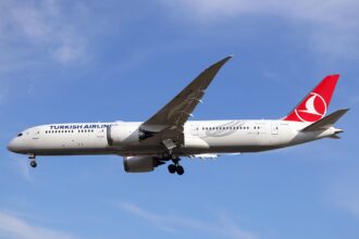 A Turkish Airlines B787-9 plane mid flight. The plane is decorated in white with red accents on its tail. The background features a blue sky with wispy clouds.