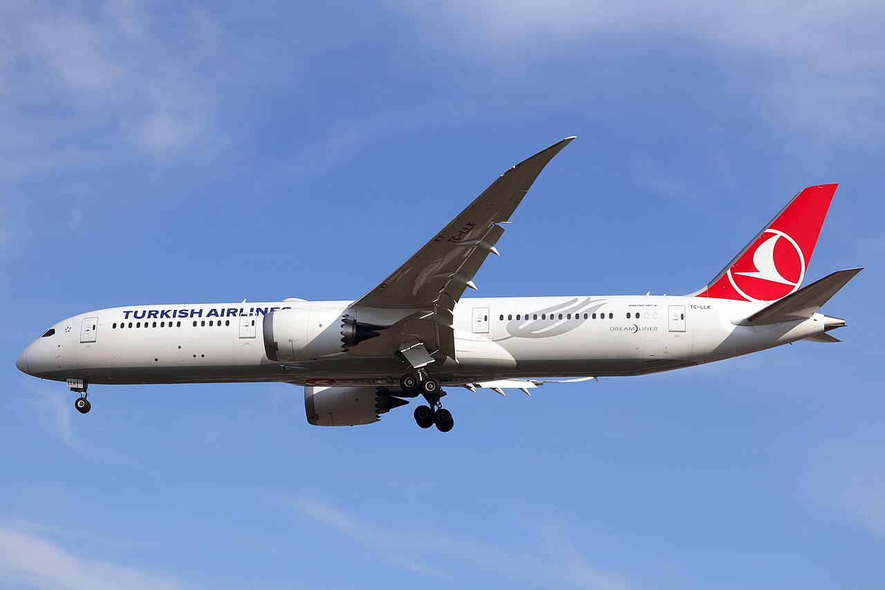 A Turkish Airlines B787-9 plane mid flight. The plane is decorated in white with red accents on its tail. The background features a blue sky with wispy clouds. These aircraft will be used in the new route development this upcoming winter.