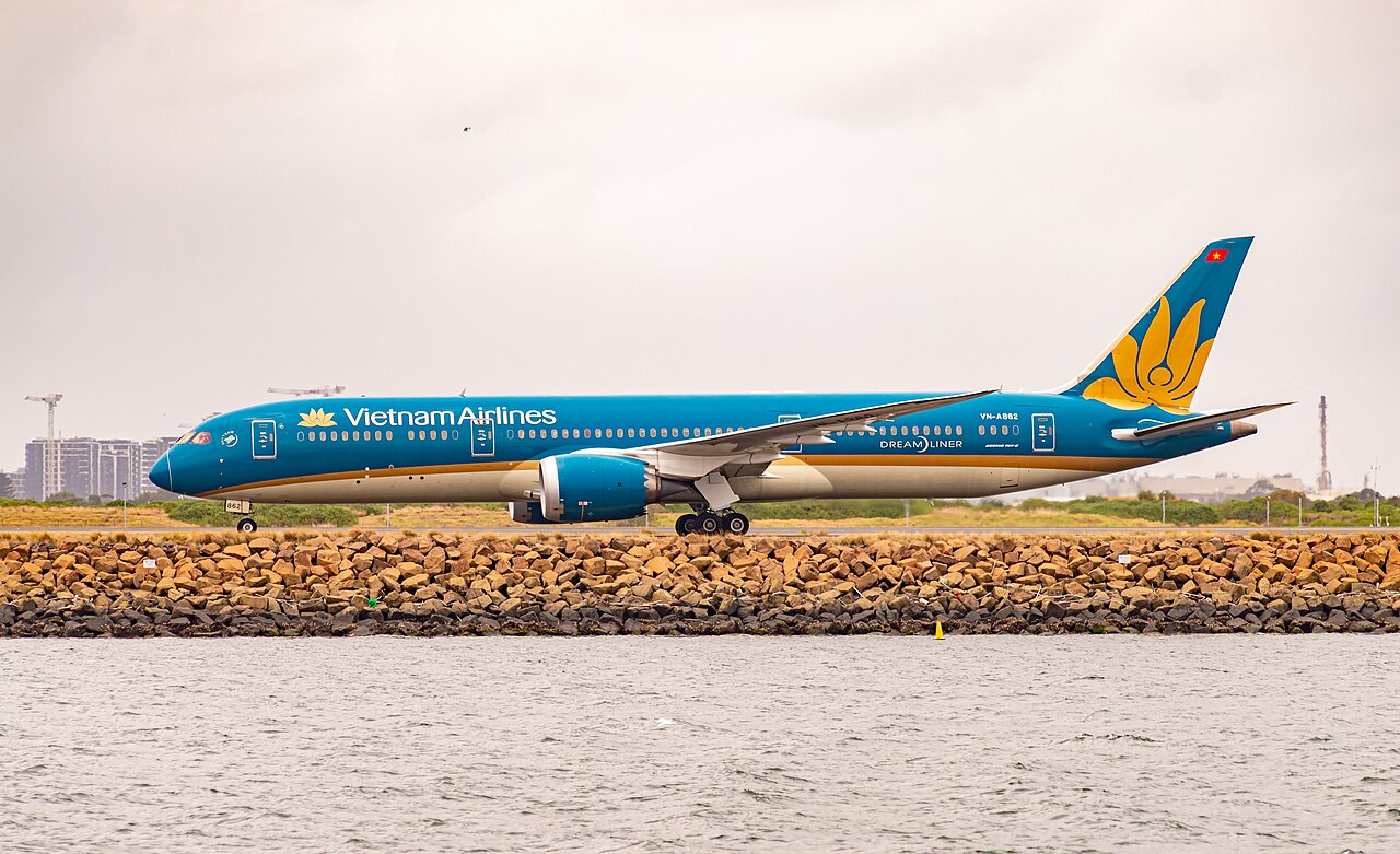 A Vietnam Airlines Boeing 787-9 Dreamliner on the runway preparing to take off. In the background is a grey and cloudy day. There's a lake positioned in front of the aircraft, whose nose is pointed to the side.