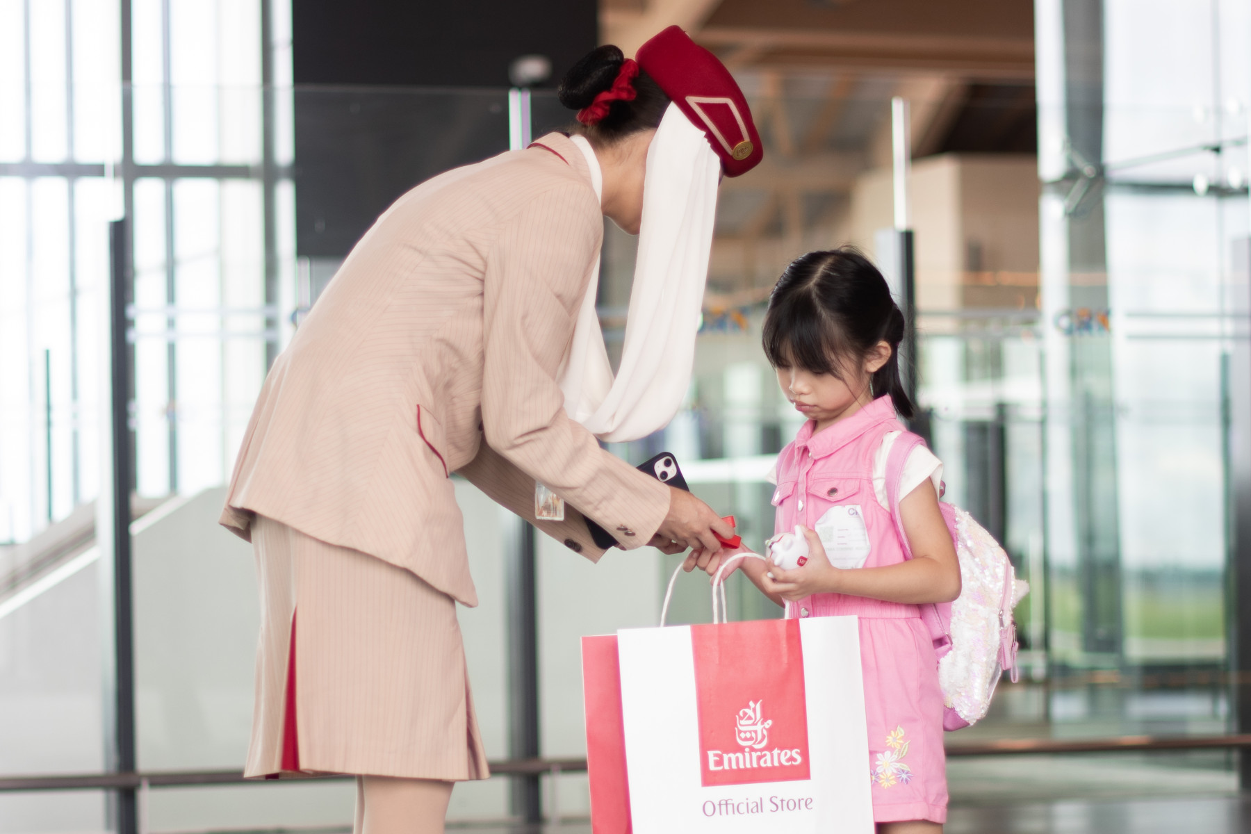 Airline cabin crew member in an Emirates uniform handing a branded shopping bag to a child with a backpack inside an airport terminal