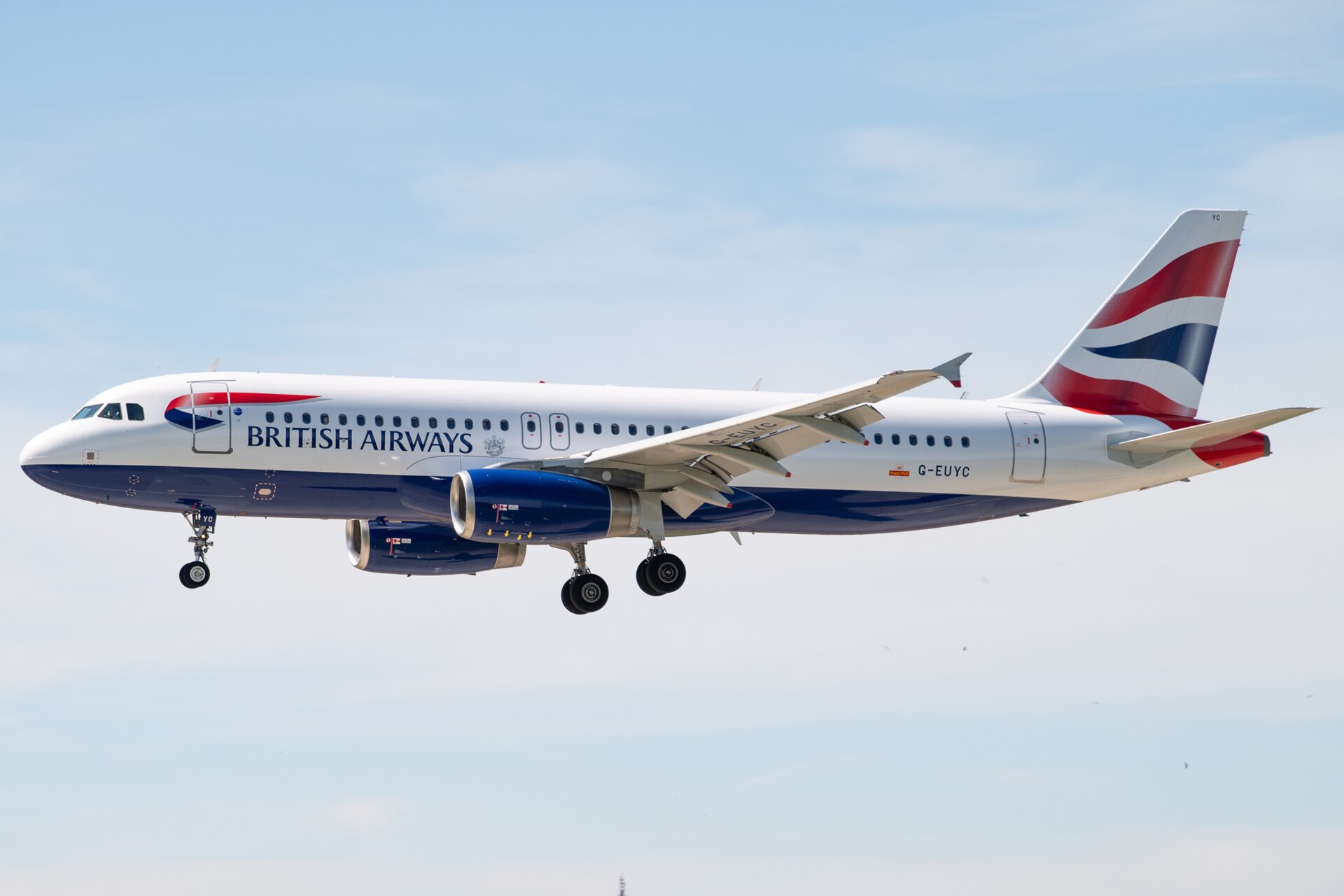 A British Airways aircraft is pictured in flight. It is pictured straight on from the side, with the nose on the centre left and the tail on the centre right. The plane is mostly white with a navy blue underbelly, blue branding along the fuselage, and a red white and blue British flag on the tail. The background is a pale blue sky with some clouds.