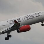 The front section of a Virgin Atlantic plane is ascending, nose to the upper right in flight. The aircraft has the airline's branding across the white fuselage, as well as red engines, and is pictured against a cloudy grey sky in the background.